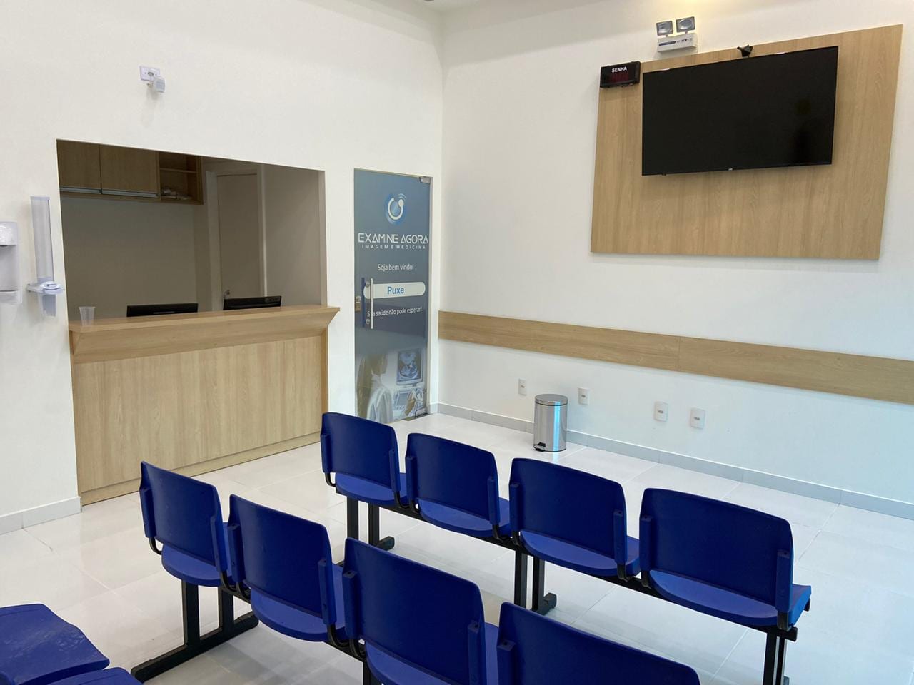 Sala de espera da clínica, com balcão de recepção em madeira clara, painel de senha e cadeiras azuis